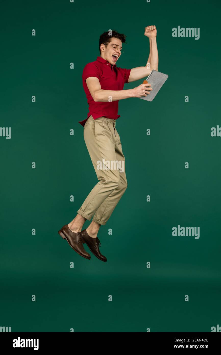 Saltando in alto, vincitore. Bel ritratto di uomo caucasico su sfondo verde studio con copyspace. Modello maschio con tablet. Concetto di emozioni umane, espressione facciale, vendite, pubblicità, moda. Foto Stock