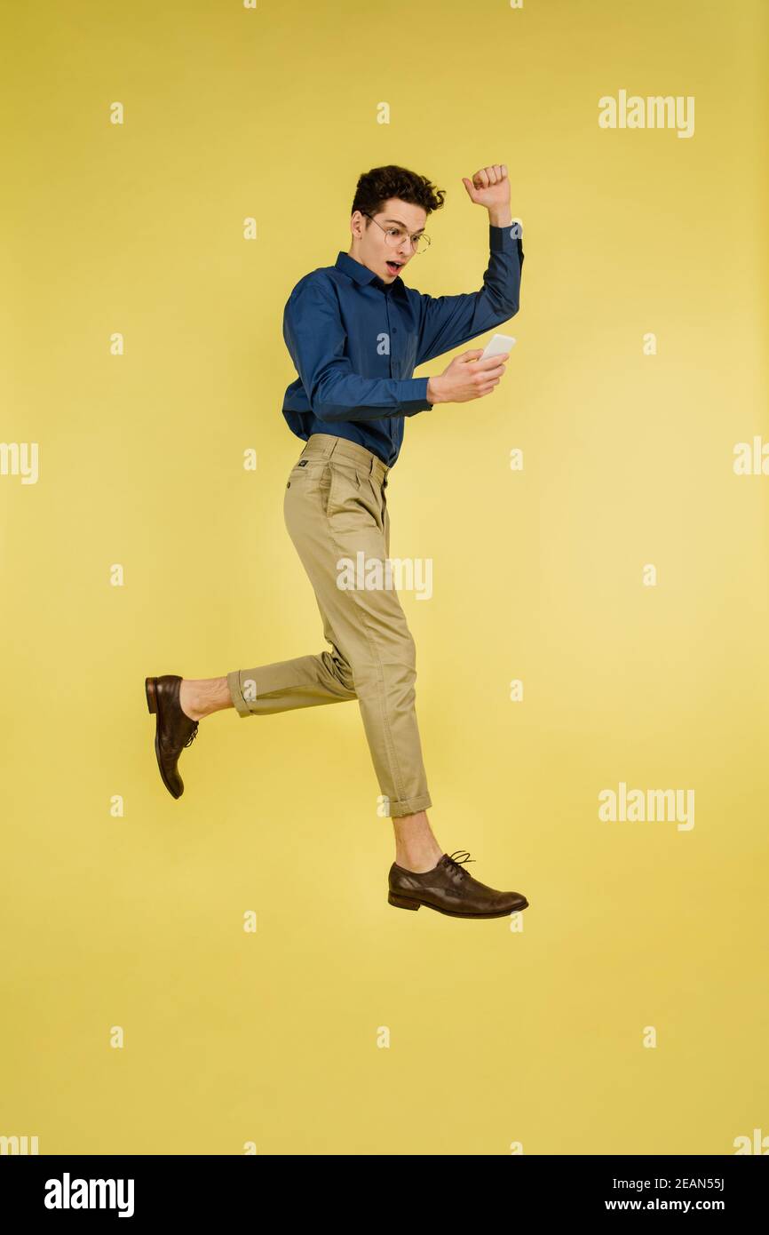 Vincitore, salto. Bel ritratto di uomo caucasico isolato su sfondo giallo studio con copyspace. Modello maschile con telefono. Concetto di emozioni umane, espressione facciale, vendite, pubblicità, moda. Foto Stock