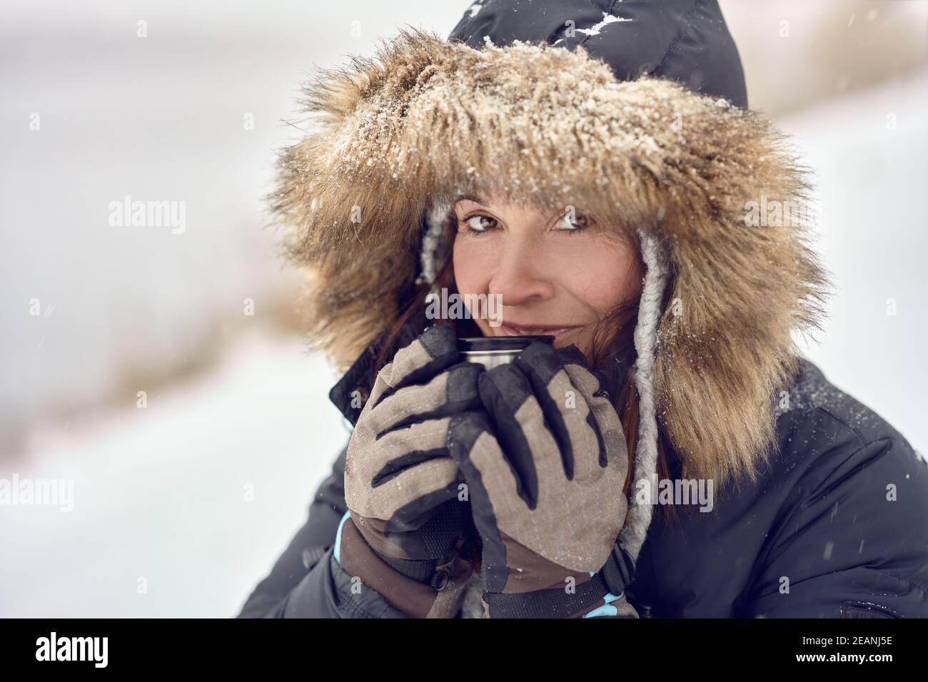 Felice donna sorridente che indossa una giacca con cappuccio rifinita in pelliccia godendo una tazza di caffè si è cullata nelle sue mani guanto all'aperto nella neve d'inverno in un concetto di t Foto Stock