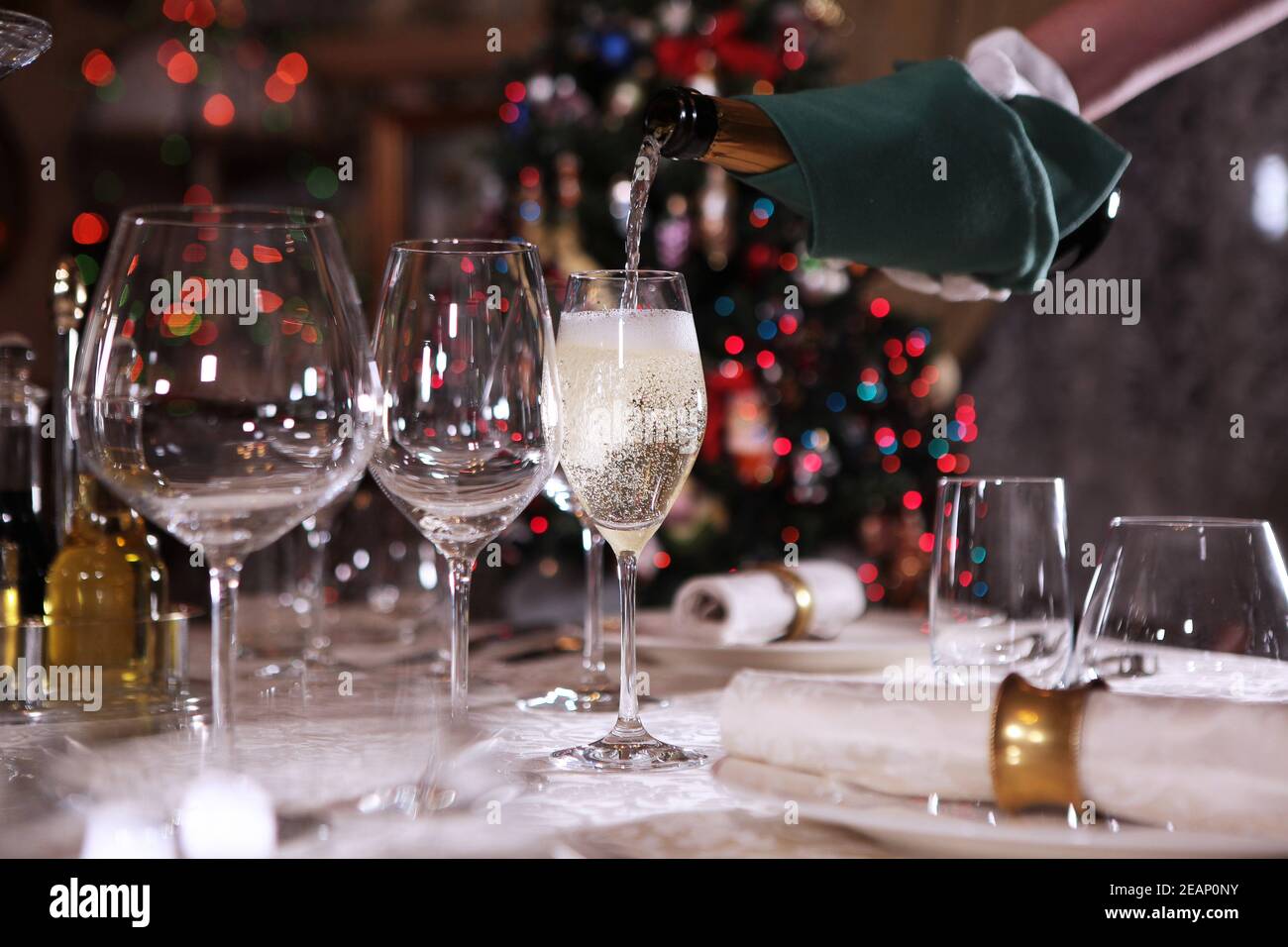 Lo champagne viene versato in un bicchiere sullo sfondo di un albero di Natale. Vino frizzante. Cameriere non riconoscibile. Impostazione del tavolo festivo. Foto Stock