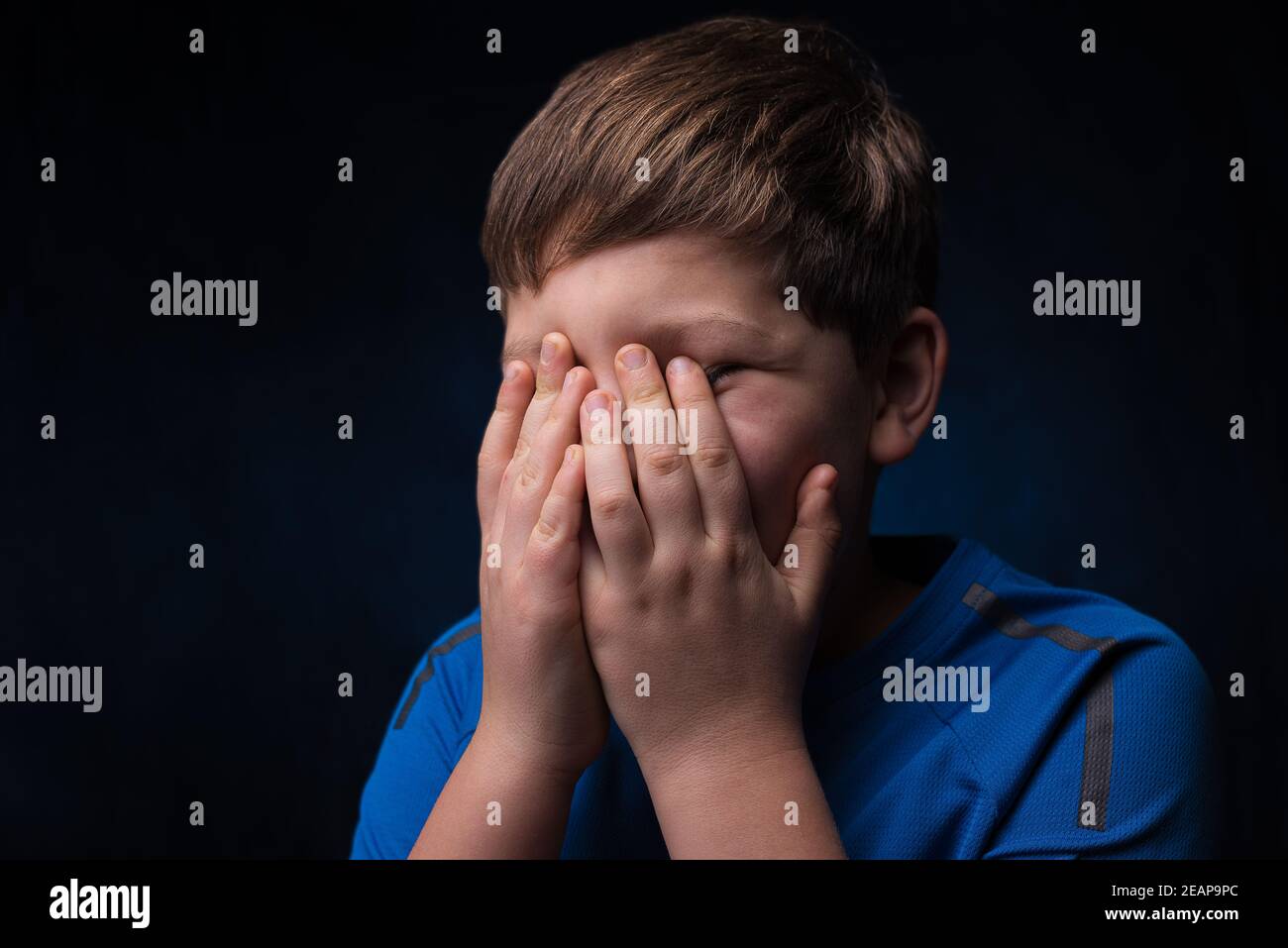 ragazzo bianco con capelli marrone chiaro in un blu t-shirt sportiva che ricopre il viso con due mani isolate sfondo Foto Stock