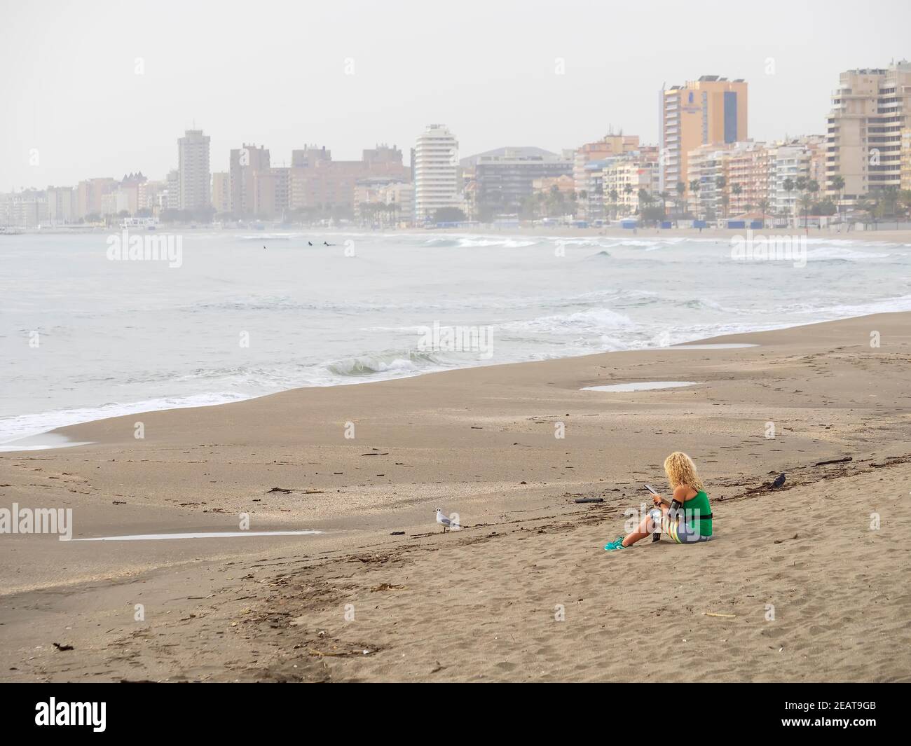 Donna seduta da sola sulla sabbia della spiaggia con il suo cellulare in mano con la città di Fuengirola sullo sfondo. Foto Stock