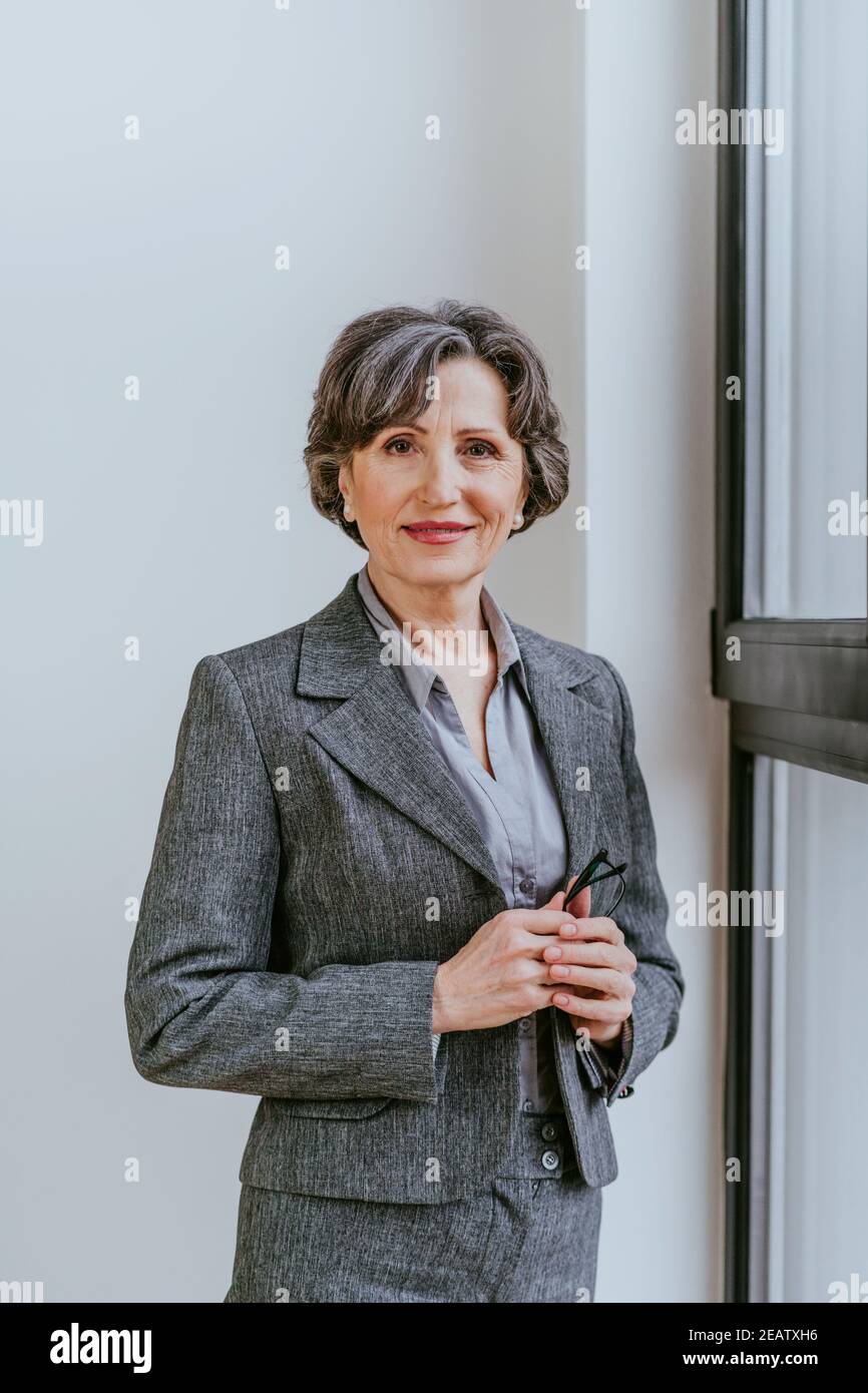 Ritratto di una donna d'affari matura che guarda la macchina fotografica in piedi in un ufficio vicino alla finestra interna. Foto Stock