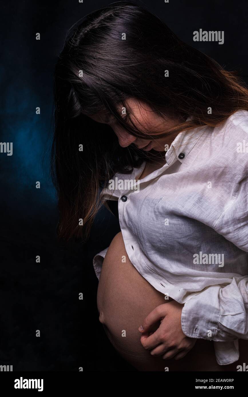 una giovane donna di gravidanza dai capelli neri in una camicia bianca con la grande età gestazionale tiene il suo grande ventre con le mani Foto Stock