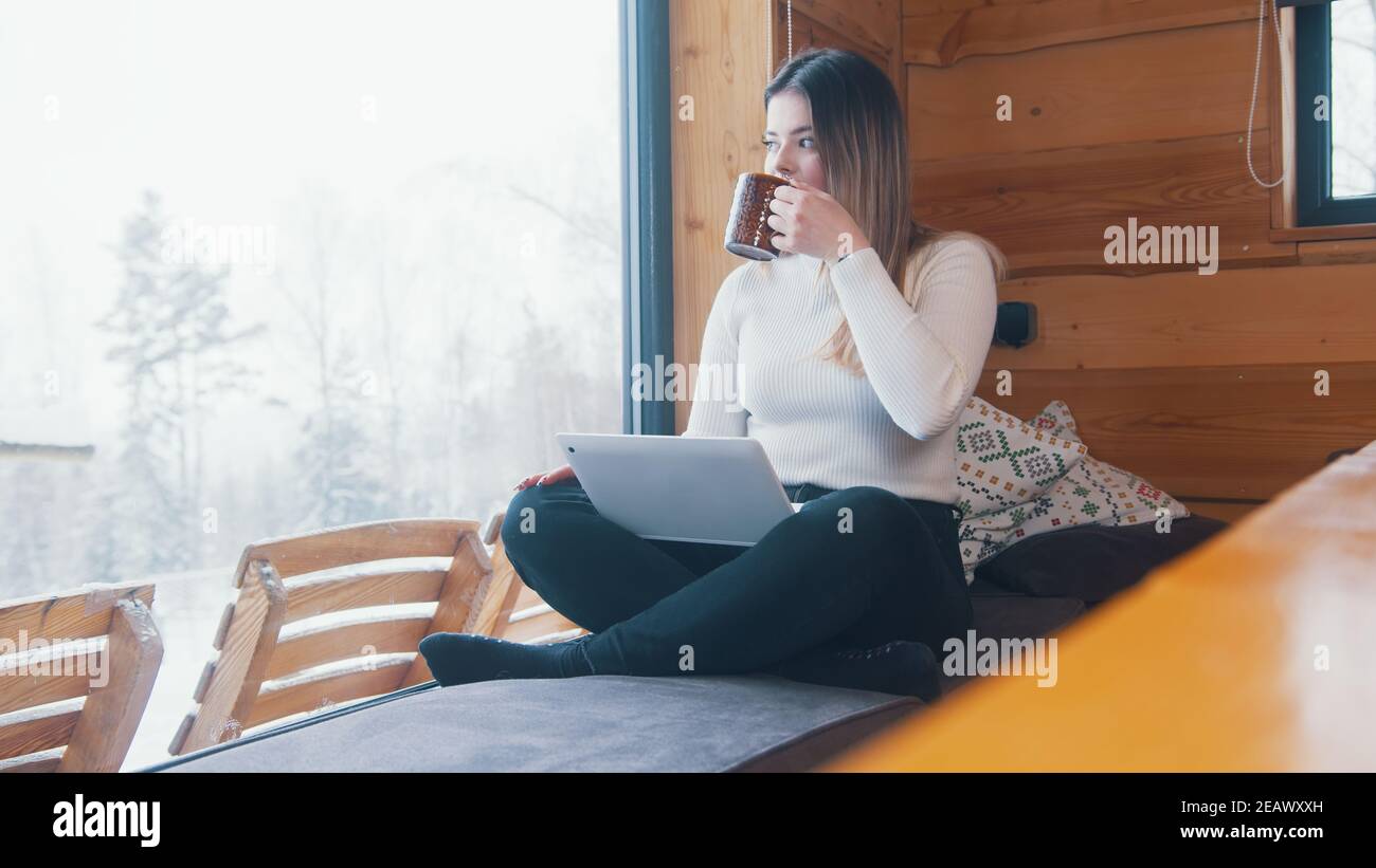 Giovane donna caucasica che beve caffè e usa un computer portatile vicino alla finestra. Vacanze invernali. Foto di alta qualità Foto Stock