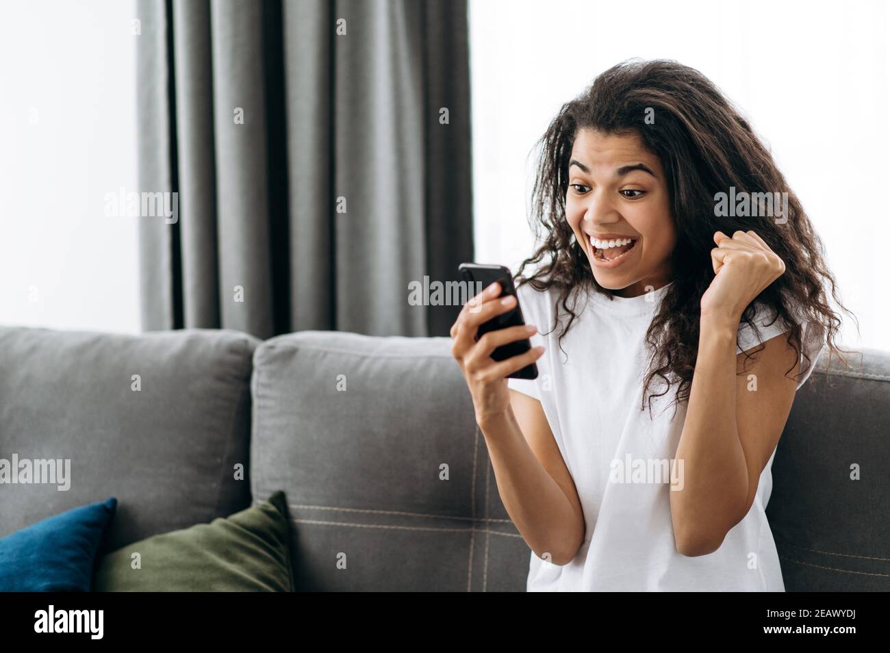 La giovane donna con grande gioia usa lo smartphone, riceve un buon messaggio. Felice afroamericana ragazza ottenere una promozione sul lavoro o superamento degli esami. Gioiosa donna libera Foto Stock