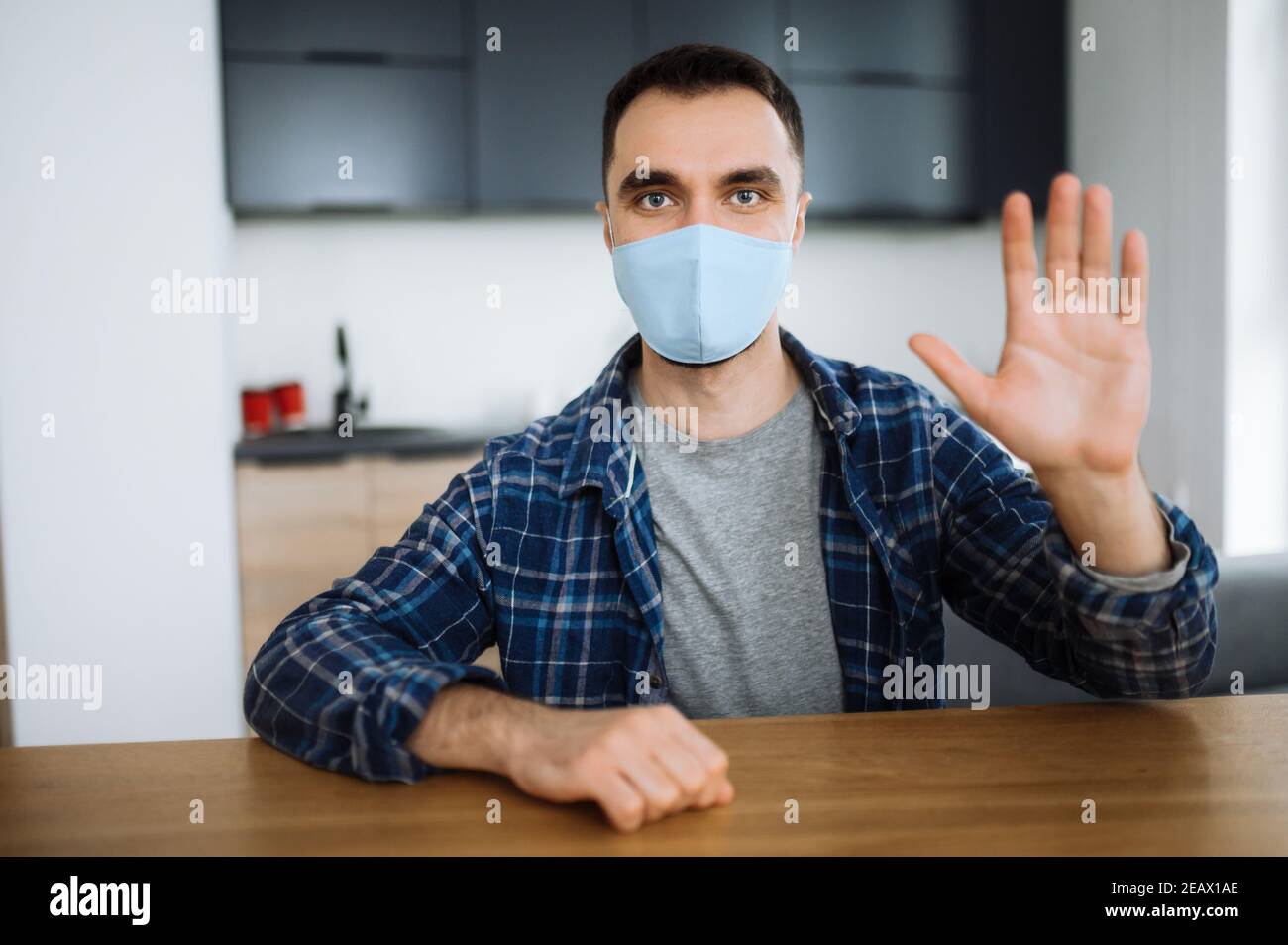 Misure preventive. Il giovane caucasico in una maschera medica protettiva si siede a casa in un abbigliamento casual, agitando la mano e guarda direttamente nella macchina fotografica Foto Stock