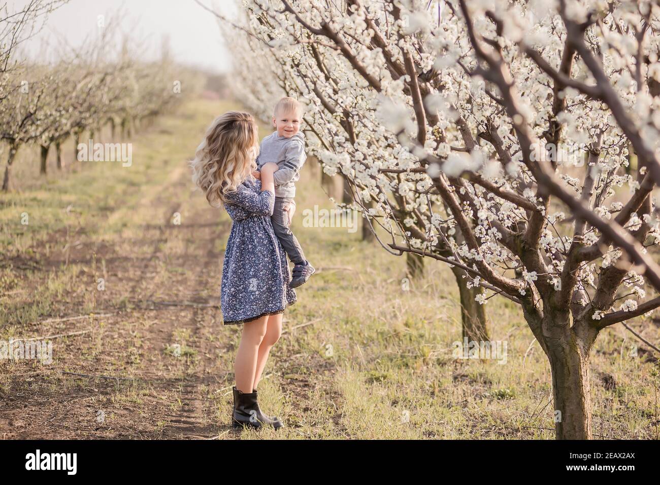 Giovane madre bionda con suo figlio cammina per mano tra i fiorenti frutteti di mele. Week-end all'aperto in un giardino con fiori bianchi. Ha Foto Stock