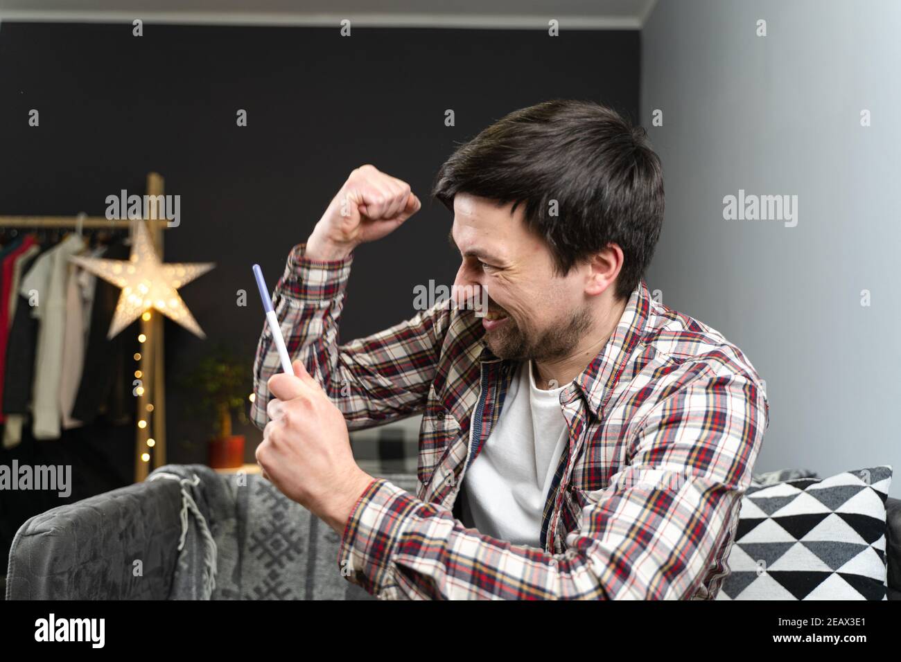 Giovane maschio caucasico felice guardando al test di gravidanza molto felice. Bell'uomo sorridente ispirato e felice e danze, canta mentre si siede sul divano Foto Stock
