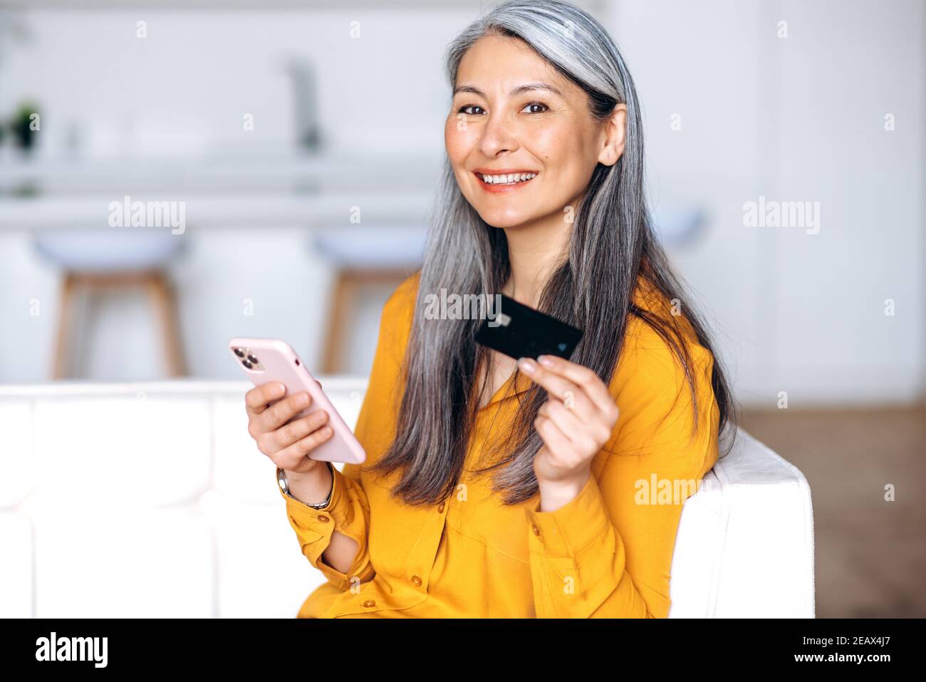 Felice metà invecchiata sorridente donna dai capelli grigi, sta facendo acquisti on-line. Gioiosa donna anziana asiatica detiene la carta di credito e il suo smartphone, lo shopping online, o Foto Stock