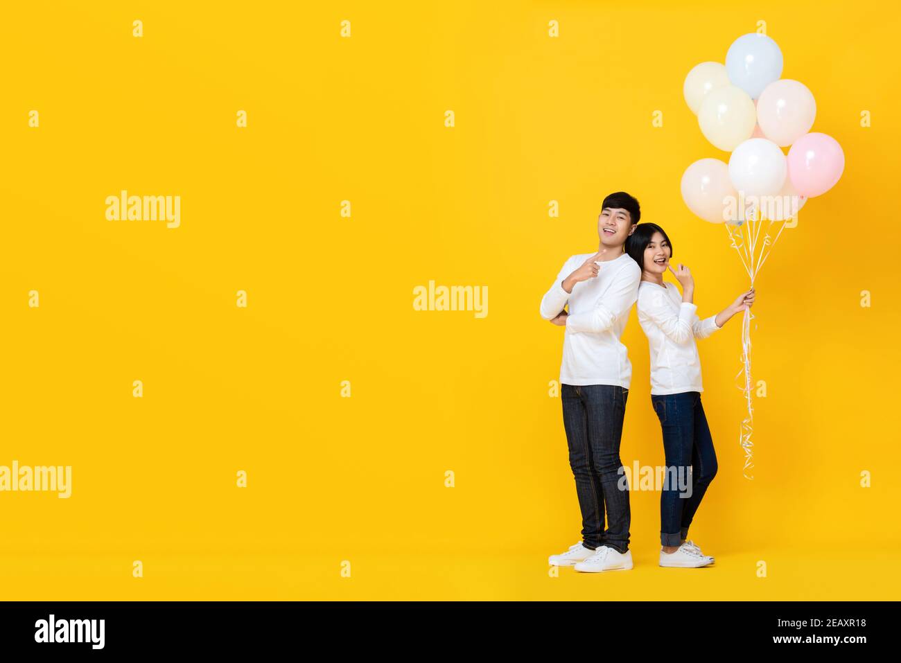 Giovane coppia asiatica allegra in piedi con sostegno back to back tenendo un mazzo di palloncini su sfondo giallo colorato Foto Stock