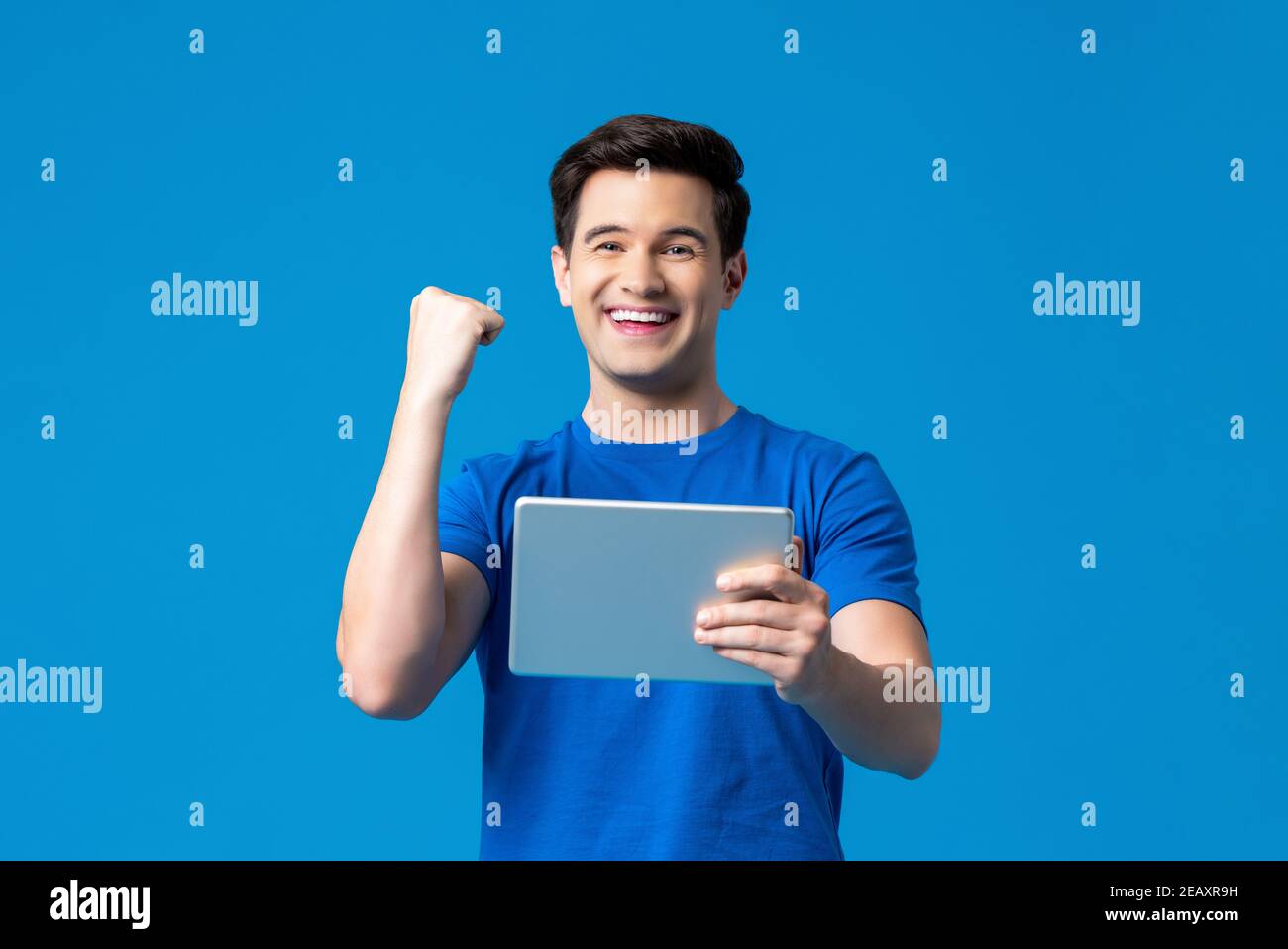 Ritratto di un uomo entusiasta che tiene il computer tablet in piedi contro il blu sfondo che acclamano con braccio rialzato Foto Stock