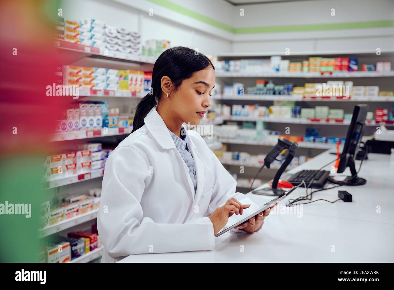 Giovane farmacista femminile in piedi dietro il banco indossando un camice da laboratorio utilizzando il digitale compressa in farmacia Foto Stock