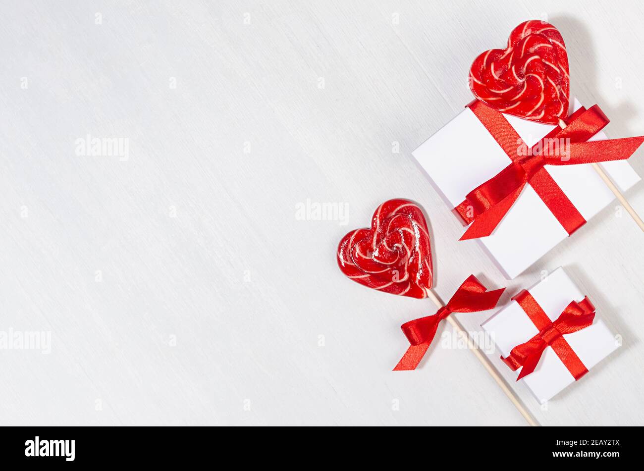 Festive Love background - scatole regalo bianche con nastro di seta rossa e cuori di lecca dolce su tavola di legno bianco, vista dall'alto, bordo. Foto Stock