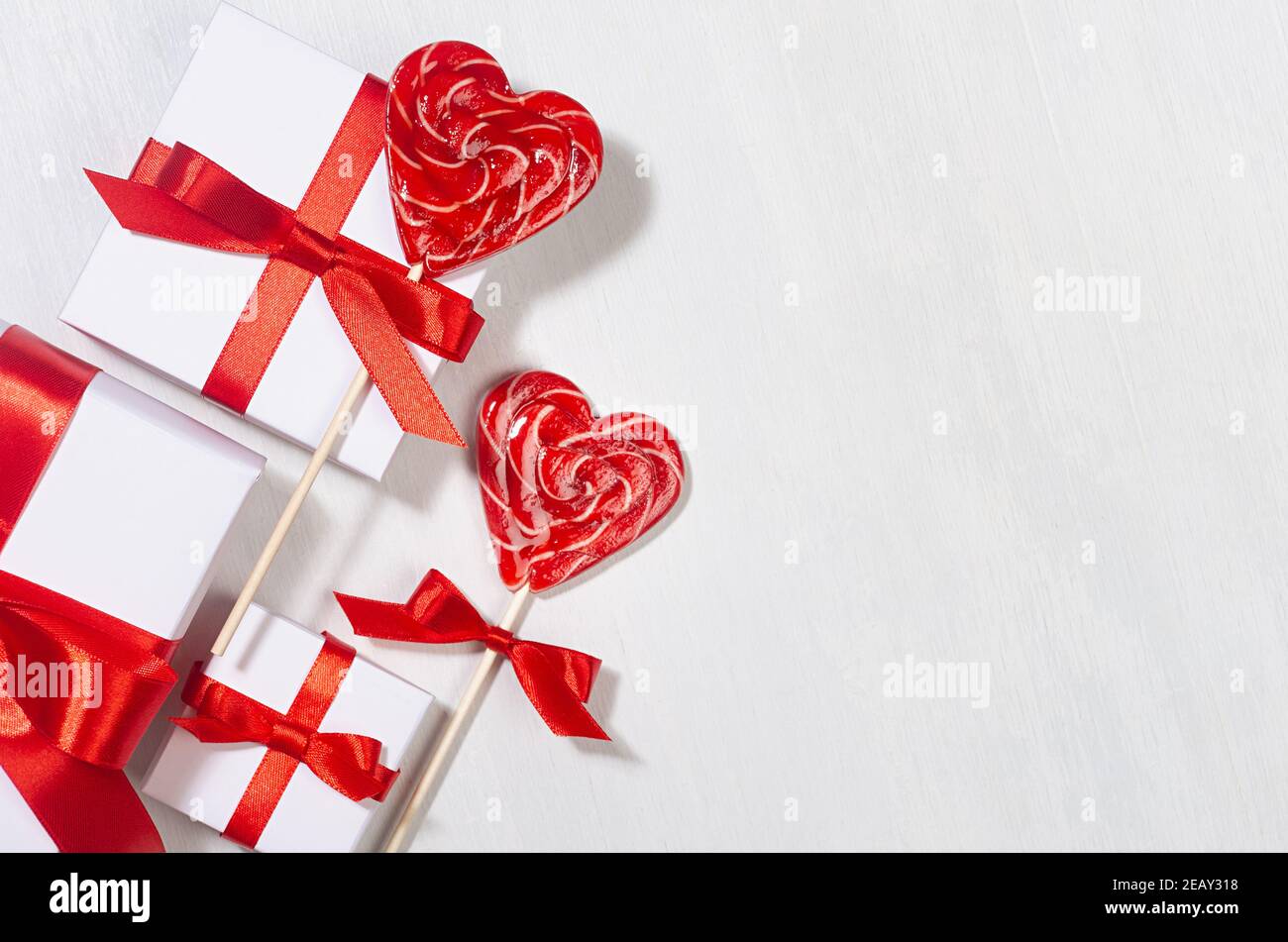 Festive Love background - scatole regalo bianche con nastro di seta rossa e cuori di lecca dolce su tavola di legno bianco, vista dall'alto, bordo. Foto Stock