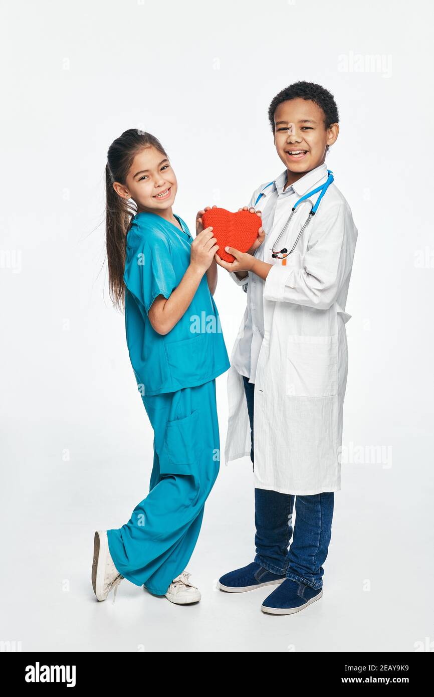 Futuri medici cardiaci. Gruppo di bambini multietnici che indossano uniformi mediche, i bambini tengono la forma del cuore nelle mani Foto Stock