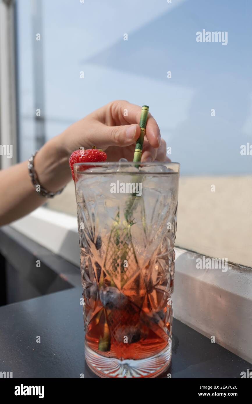 Fuoco selettivo sulla mano di una donna con bicchiere da cocktail in cui una bevanda rossa con cubetti di ghiaccio e una fragola. Lei agita il bicchiere con un foglio Foto Stock
