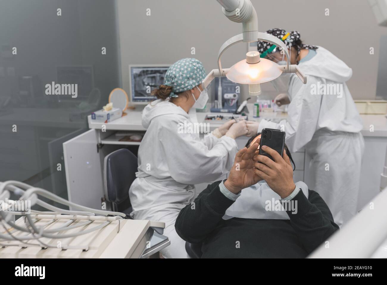 Paziente che utilizza uno smartphone in studio dentistico. Due dottoresse che preparano la procedura. Concetto di igiene e salute dentale Foto Stock