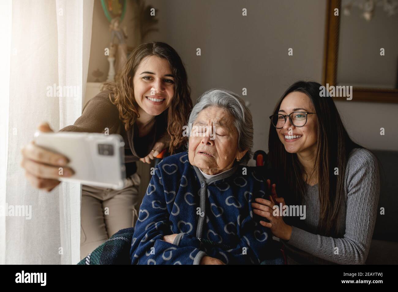 Due nipoti con la vecchia nonna in sedia a rotelle scattano una foto o fanno una videochiamata con volti sorridenti. Nuovo concetto normale, di terza età, di famiglia. Foto Stock