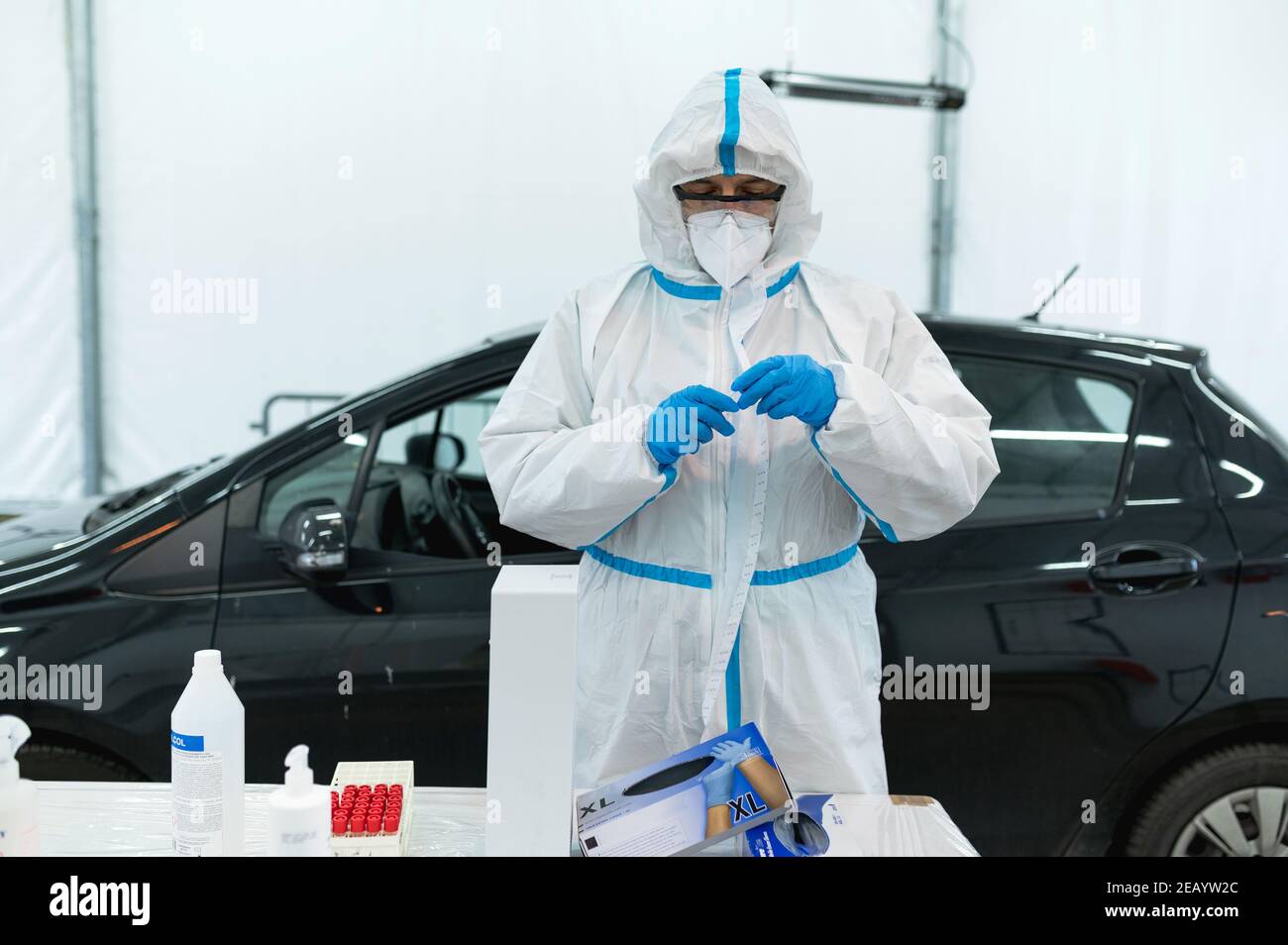 Infermiere indossare tuta protettiva e attrezzature di protezione personale si preparano a prendere bastoncino con l'auto in background. Stazione drive-thru per il test del coronavirus. Foto Stock