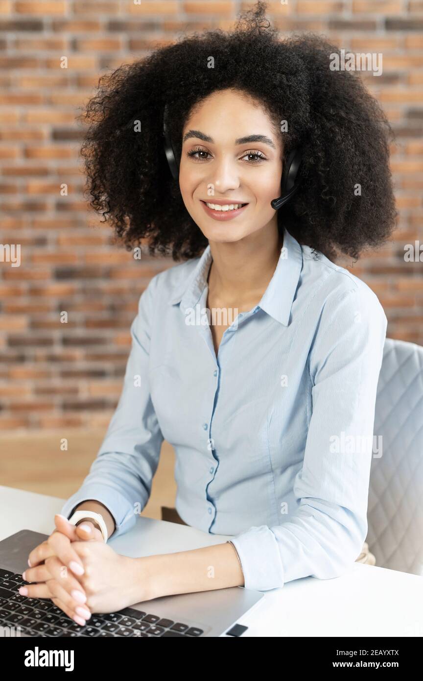 Ritratto formale di una giovane donna intelligente di razza mista con acconciatura afro in cuffia sorridente, seduto alla scrivania, lavorando nel reparto vendite Foto Stock