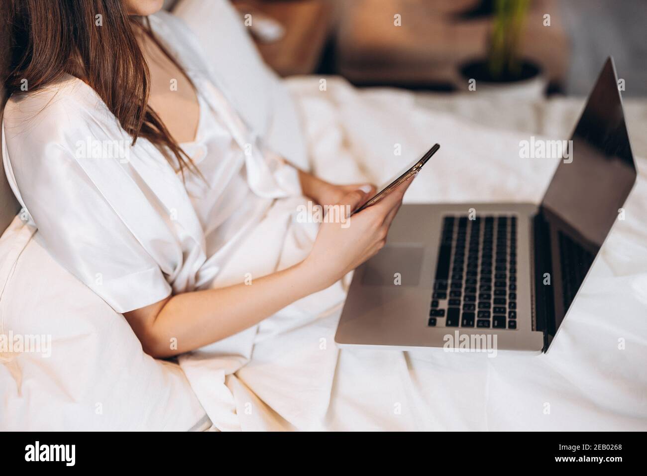 computer portatile e telefono nelle mani di una donna senza volto. computer portatile e telefono sullo sfondo di pigiami bianchi. lavoro a casa Foto Stock