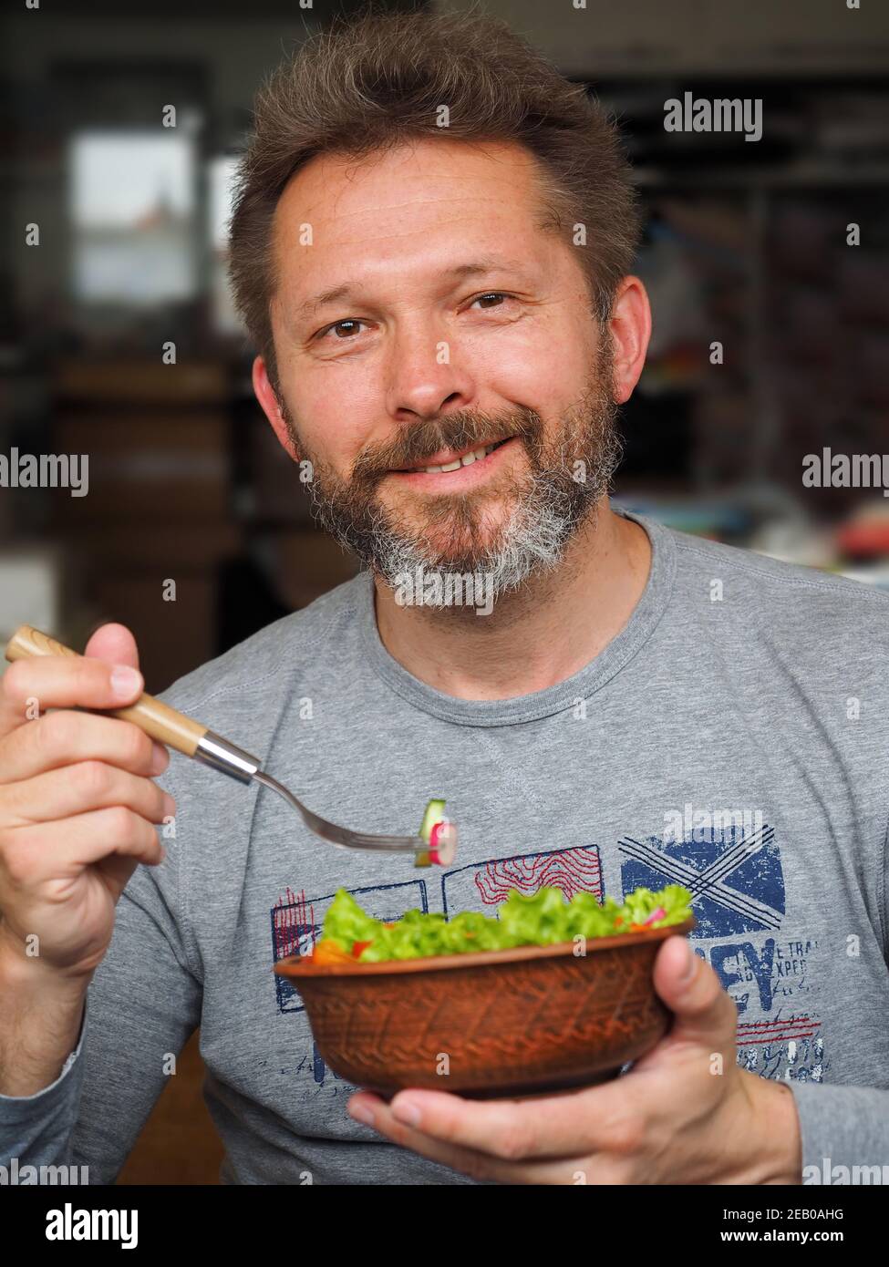 Sorridente uomo di 35-45 anni con barba mangia una deliziosa insalata verde con pomodori d'arancia, ravanelli e cetrioli. Foto Stock