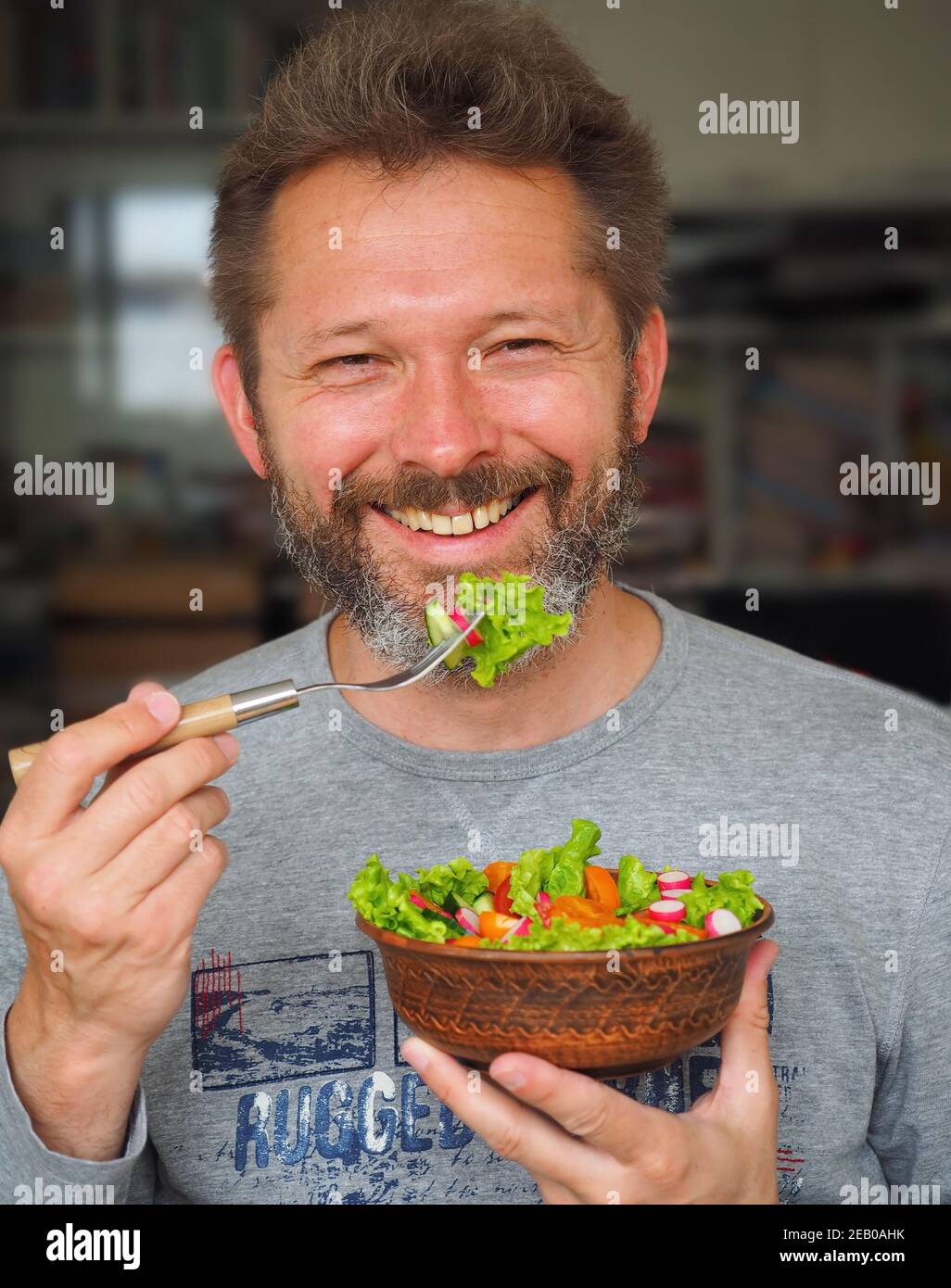 Sorridente uomo di 35-45 anni con barba mangia una deliziosa insalata verde con pomodori d'arancia, ravanelli e cetrioli. Foto Stock