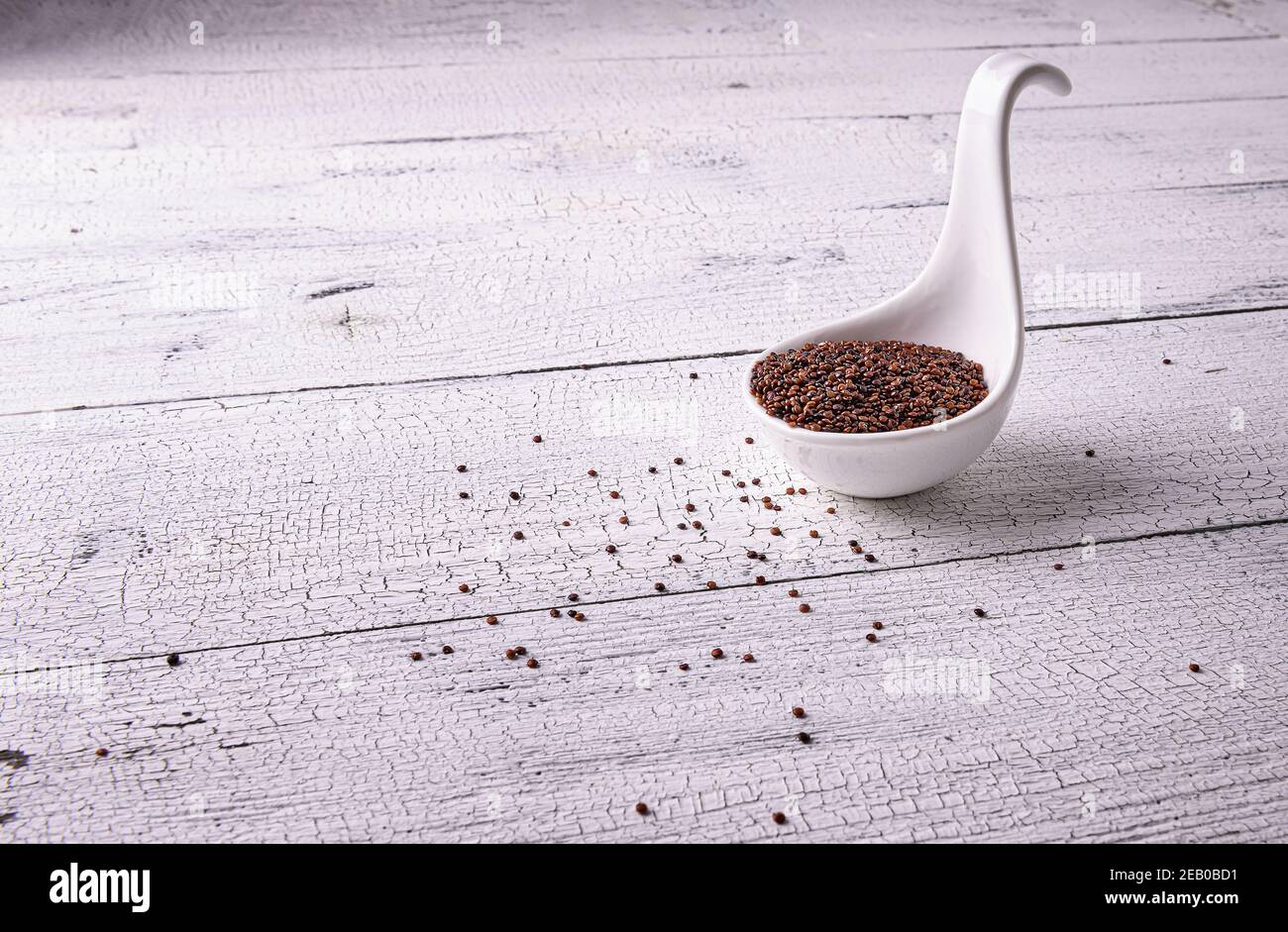 Mucchio di semi di quinoa grezzi rossi in ciotola a cucchiaio bianca su tavola di legno verniciato a trama bianca. Pochi semi sono sparsi intorno. Cibo eccellente con copia Foto Stock