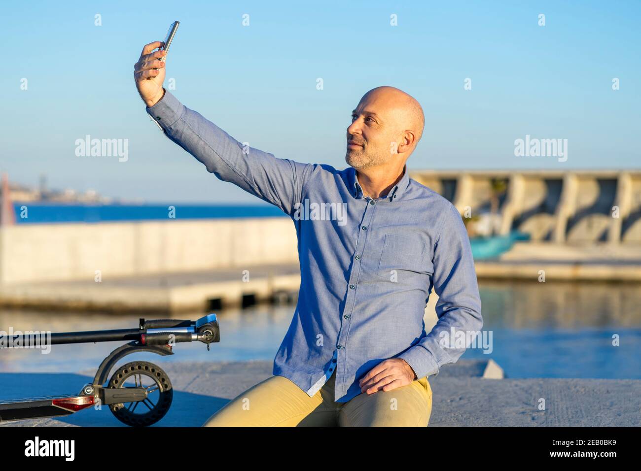 L'uomo maturo prende selfie con il telefono cellulare. È seduto e ha uno scooter elettrico. Sullo sfondo si può vedere il mare. C'è la luce del tramonto. Ho Foto Stock