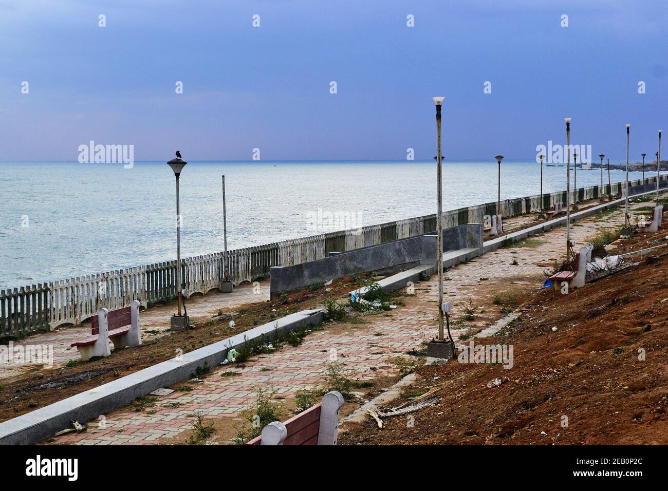 Passerella sul lungomare sporca danneggiata con panca rotta e luci di strada. Cielo nuvoloso sovrastato sul mare. Kanyakumari, Tamil Nadu, India. Foto Stock