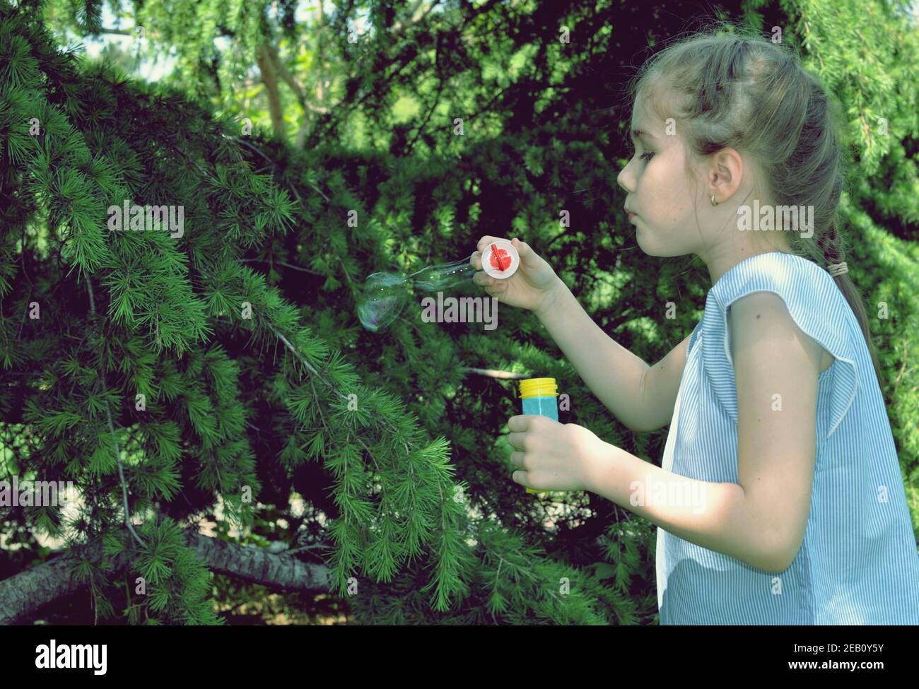 Ritratto laterale di una bella bambina bionda che soffia bolle, con alberi verdi sullo sfondo del parco. Spazio di copia, messa a fuoco selettiva Foto Stock