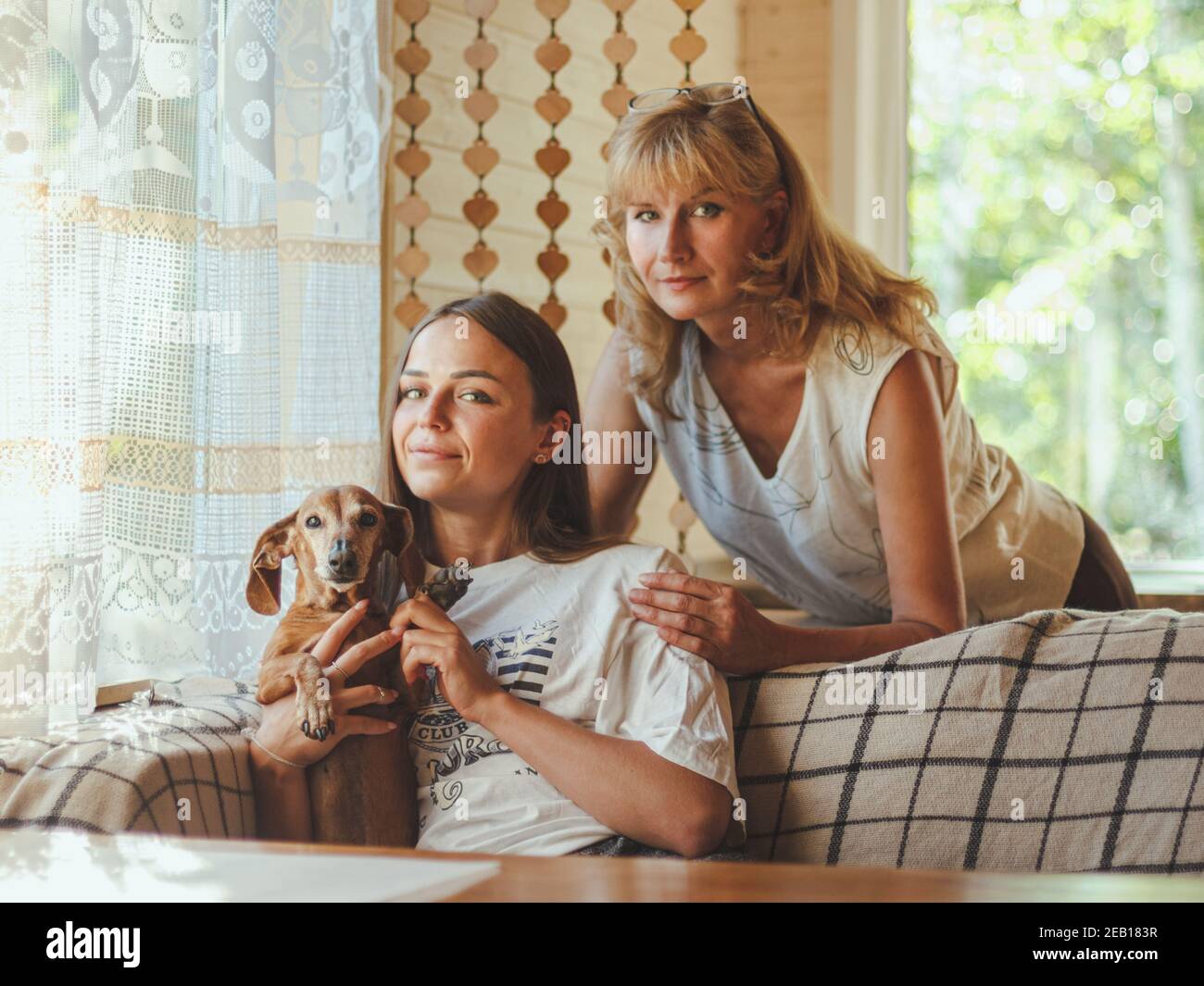 Amorevole anziana madre abbraccio adulto 20s figlia da dietro mentre figlia seduta sul divano e tenendo il loro cane. Guardando la macchina fotografica, sorridendo e sente h Foto Stock