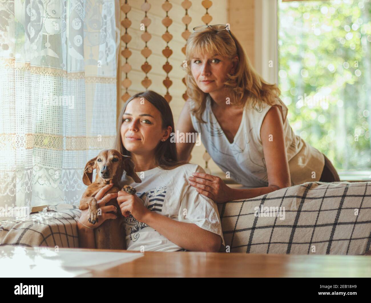 Amorevole anziana madre abbraccio adulto 20s figlia da dietro mentre figlia seduta sul divano e tenendo il loro cane. Guardando la macchina fotografica, sorridendo e sente h Foto Stock