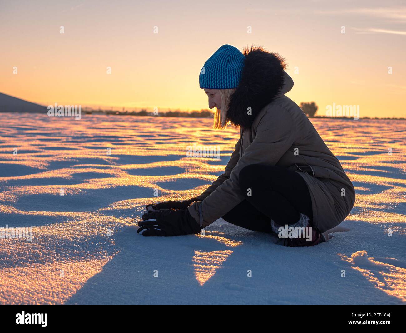 bella foto di una donna accovacciata giù raccogliendo la neve dentro un bellissimo campo di neve al tramonto Foto Stock