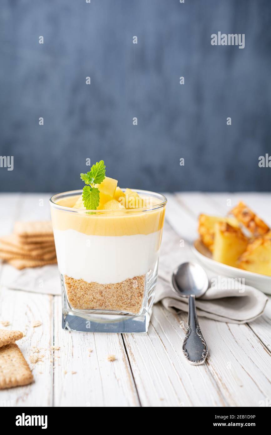 Dolce dessert a strati con cracker di graham schiacciati, formaggio cremoso e cagliata di ananas, condito con fette di frutta fresca in un vaso di vetro su vecchio dorso di legno Foto Stock