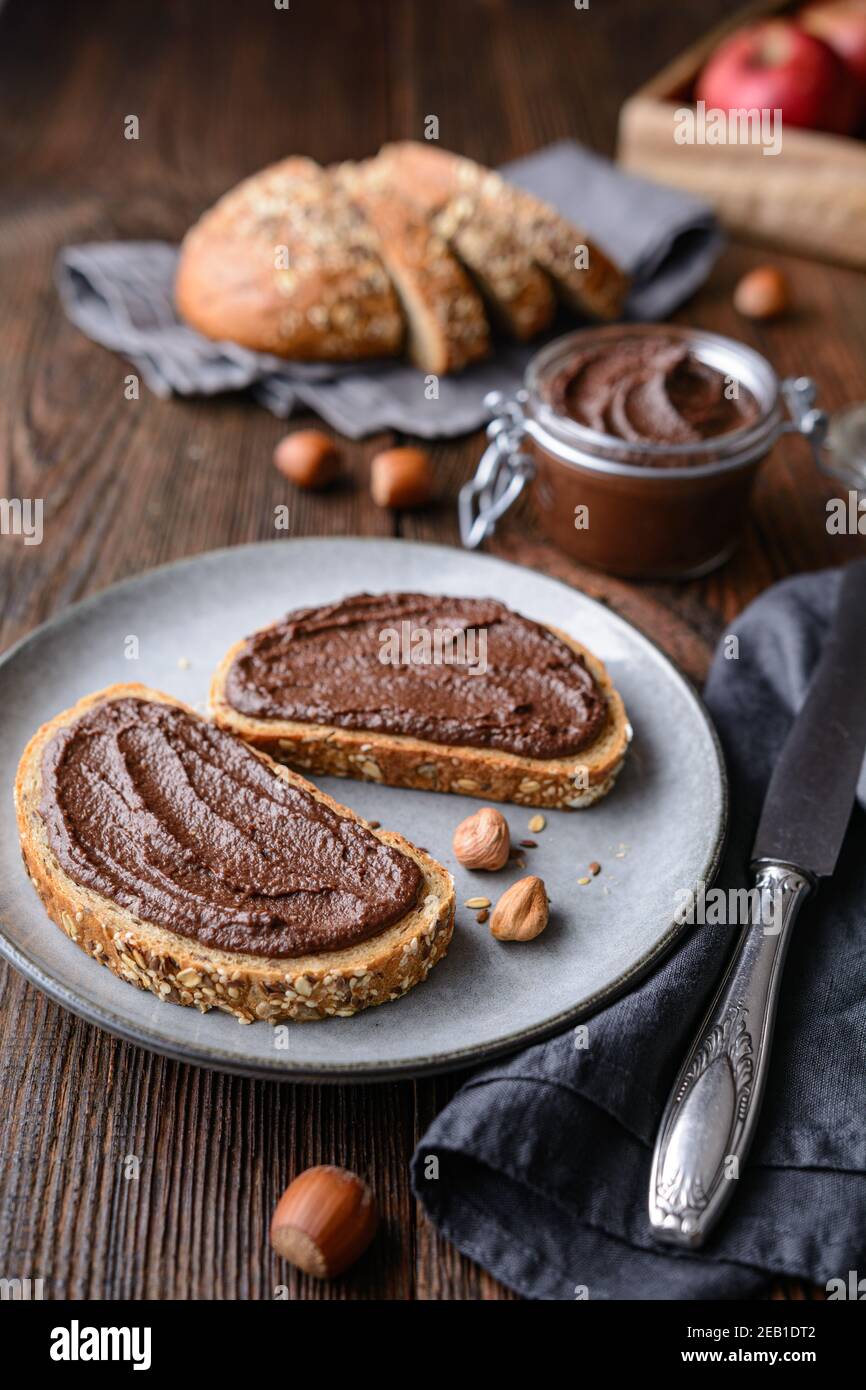 Colazione sana, fette di pane integrale con mela spalmata di cacao in polvere, nocciole tostate macinate e cioccolato fondente su legno rustico Foto Stock