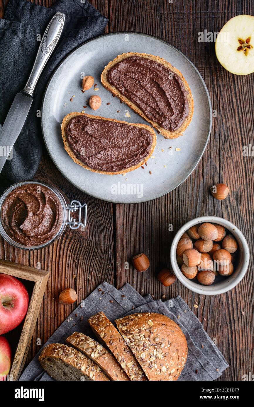 Colazione sana, fette di pane integrale con mela spalmata di cacao in polvere, nocciole tostate macinate e cioccolato fondente su legno rustico Foto Stock