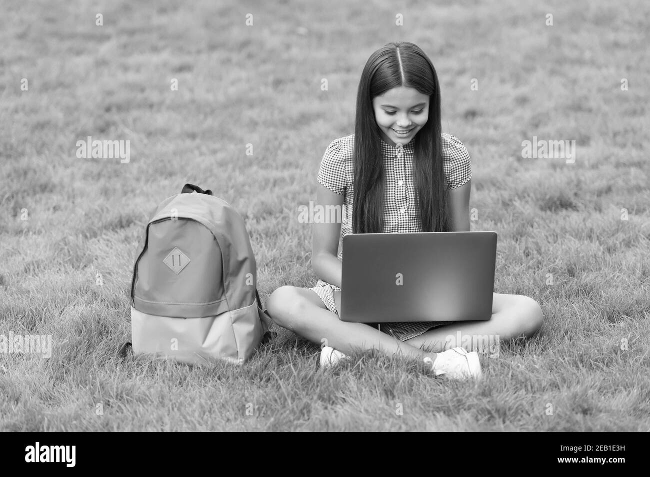 bambino felice che usa il laptop per l'istruzione scolastica online seduto in parco su erba verde con zaino, infanzia. Foto Stock