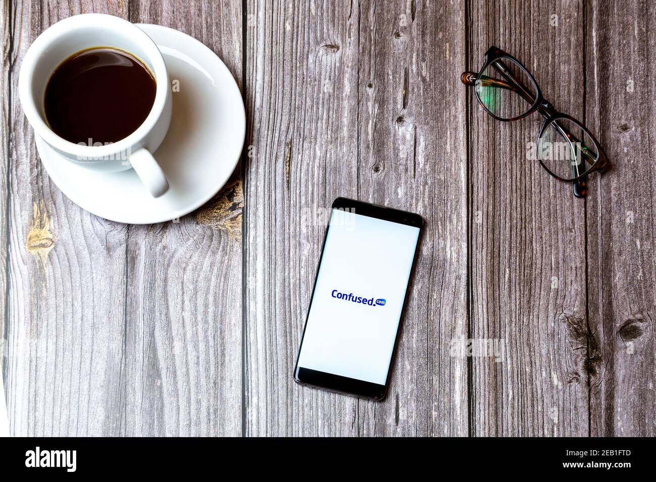 Un telefono cellulare o un telefono cellulare posato su un legno Tabella con l'app Confused.com aperta sullo schermo accanto a. un caffè Foto Stock