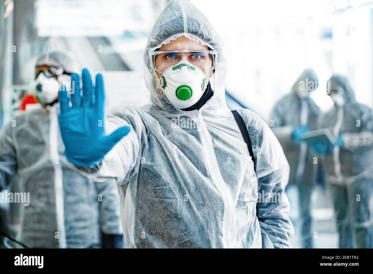 Operatore sanitario che mostra il gesto di arresto. Un team di operatori sanitari che indossano tute nocciole che lavorano insieme nel centro commerciale, per controllare un focolaio di Foto Stock