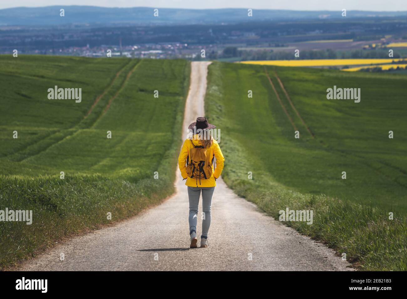 Lungo viaggio. Donna escursionista a piedi su strada vuota. Escursionista con cappello e zaino con giacca impermeabile gialla. Concetto di viaggio. Allontanarsi da esso un Foto Stock