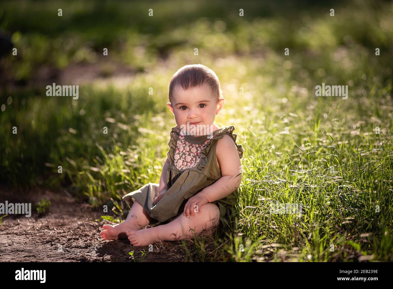 Un piccolo bambino carino con i capelli corti, in un vestito verde si siede sul prato in erba vicino al sentiero, guarda in su nel cielo al sole. Lei sorride a lei Foto Stock