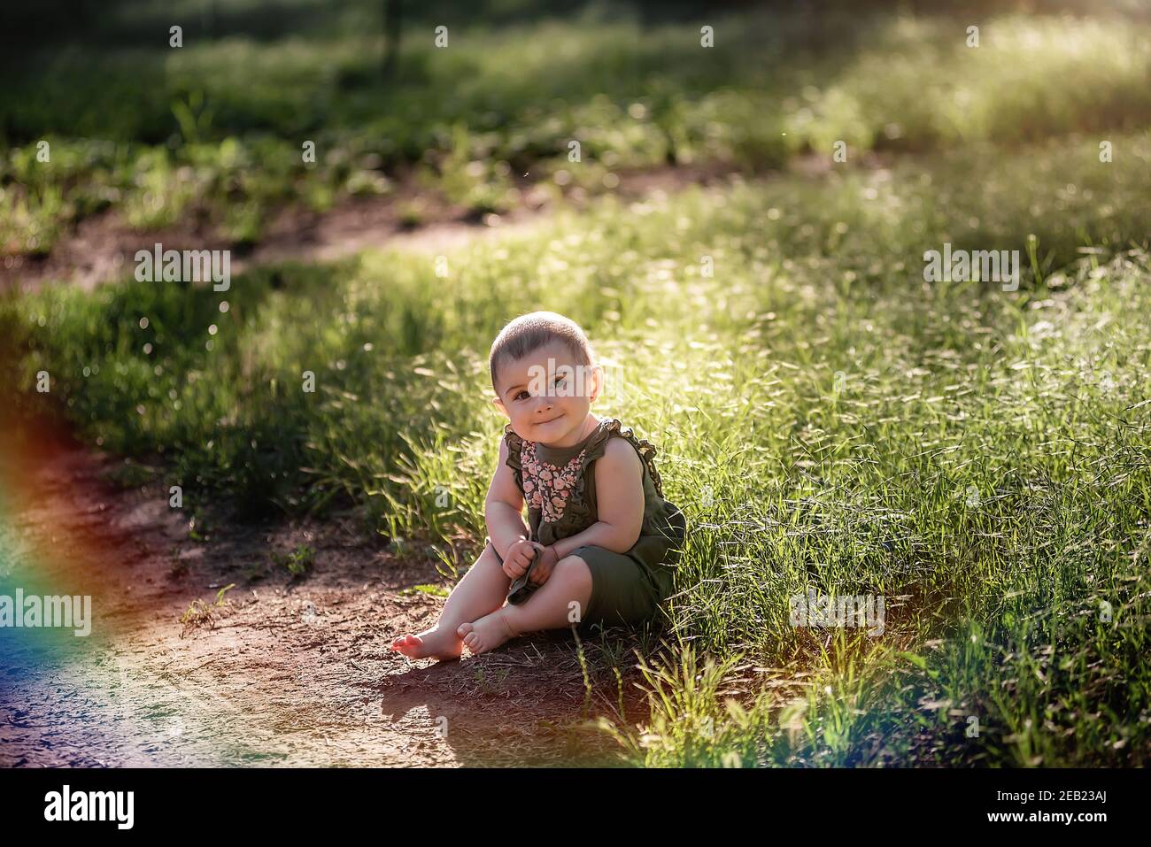 Un piccolo bambino carino con i capelli corti, in un vestito verde si siede sul prato in erba vicino al sentiero, guarda in su nel cielo al sole. Lei sorride a lei Foto Stock