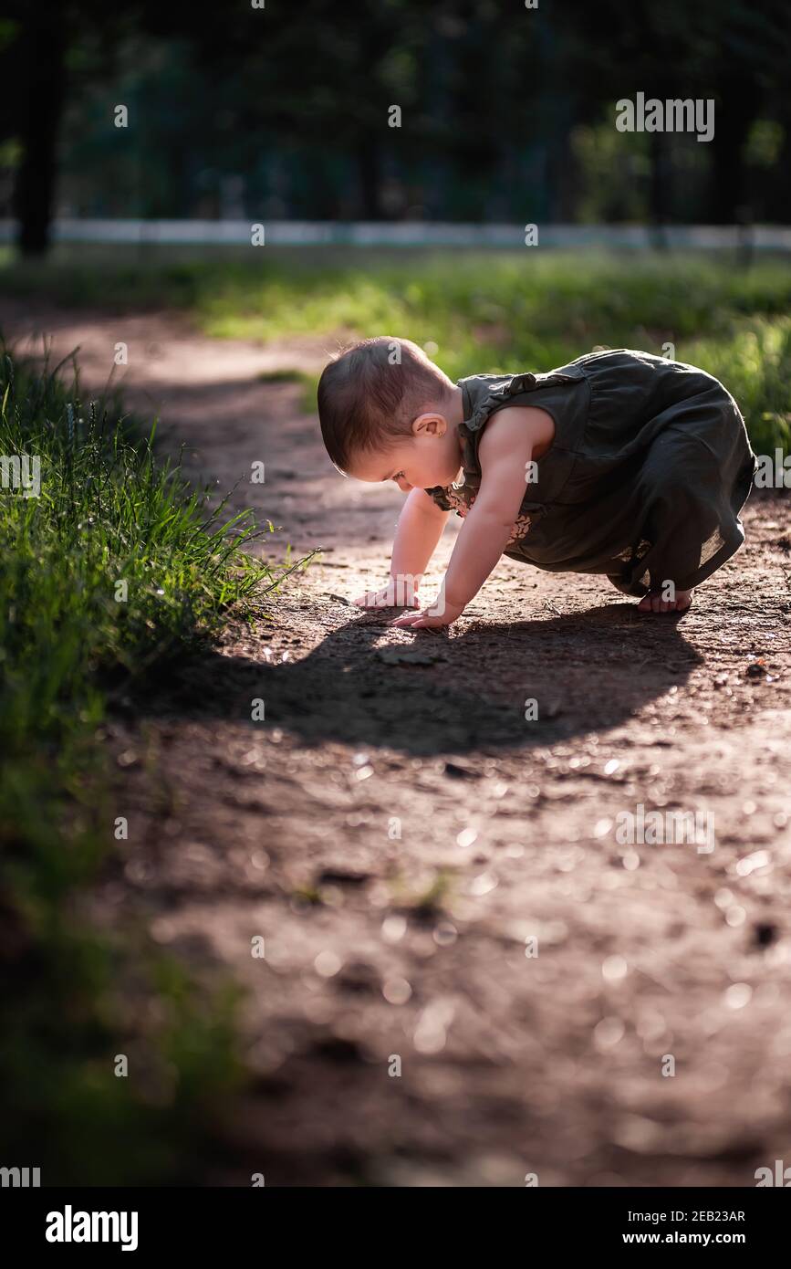 Bambino in un vestito verde, a piedi nudi, cercando di fare i primi passi. La bambina cammina con la sua famiglia nel parco lungo un sentiero terroso tra alberi verdi, Foto Stock
