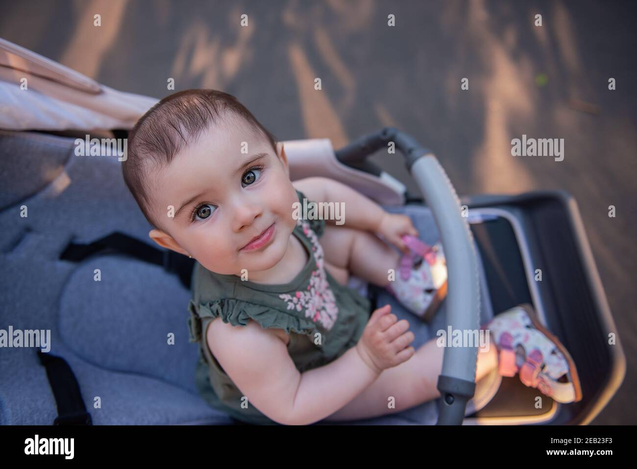 Una bambina carina con gli occhi verdi in un abito si siede in un passeggino in un parco verde. Ritratto in primo piano di un bambino che guarda nella fotocamera. Copia s Foto Stock