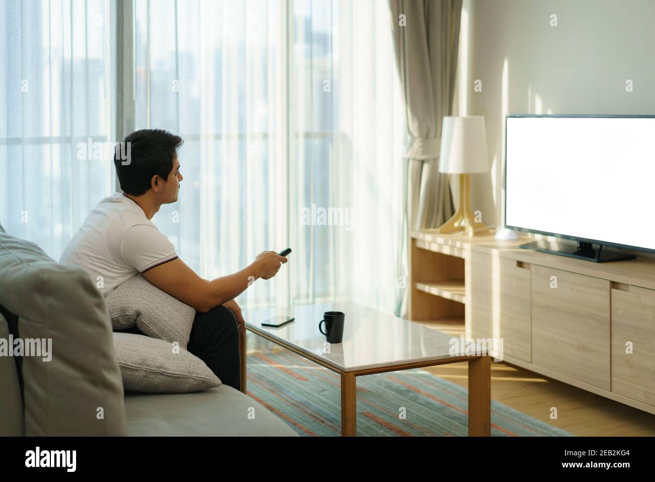 Un uomo asiatico ha un telecomando TELEVISIVO e sta premendo il canale mentre guarda la TV sul divano nel soggiorno di casa. Foto Stock