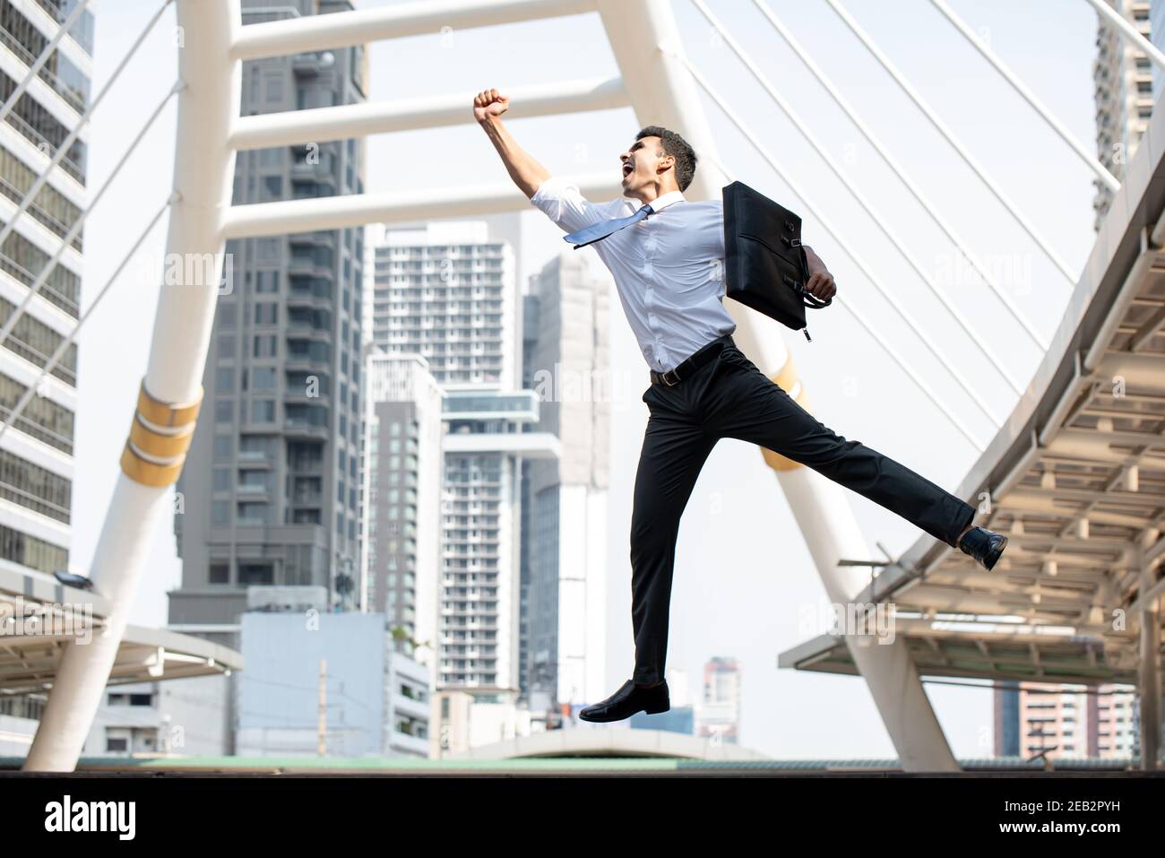 Giovane uomo d'affari asiatico felice che porta una valigetta nera che salta dentro a mezz'aria con un braccio sollevato in gesto di successo con la città edificio sullo sfondo Foto Stock