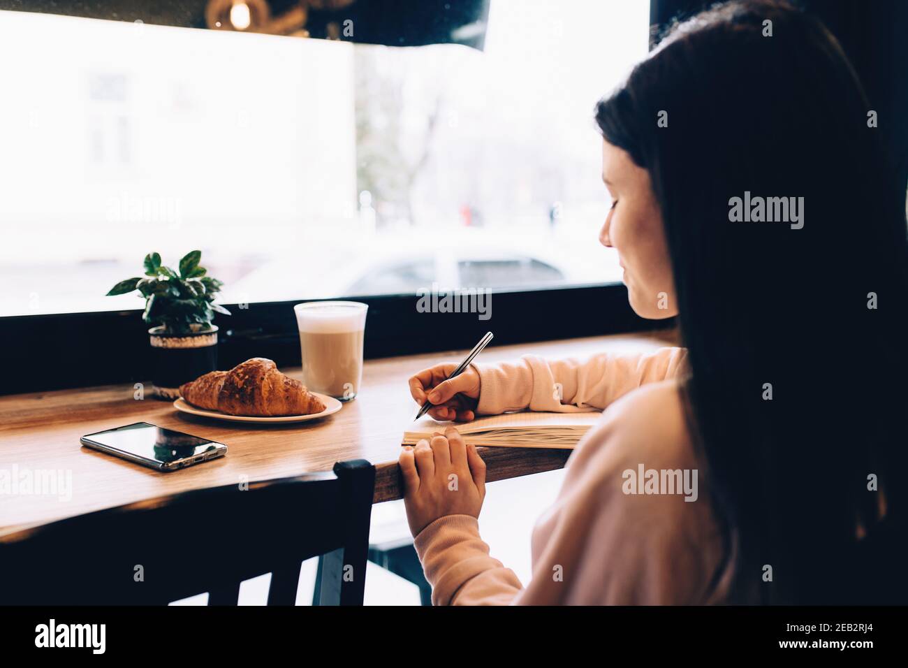 Ragazza che scrive in taccuino, croissant, caffè, telefono su tavolo di legno. Sfocato. Foto di alta qualità Foto Stock