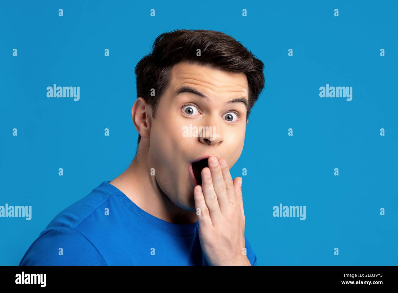 Ritratto del modello maschile caucasico in gesto sorprendente con bocca di copertura a mano su sfondo blu isolato studio, focalizzato selettivo Foto Stock